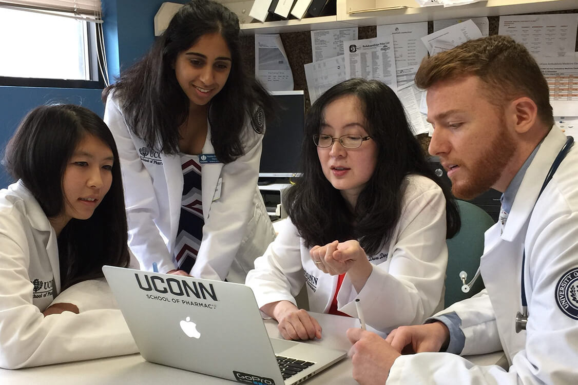 Students in whitecoats huddled around a laptop.